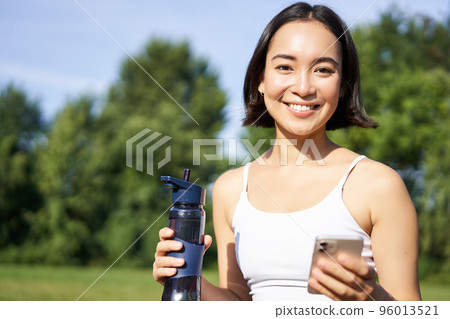 Sportswoman workout with smartphone, drinks water, holds mobile phone and smiles at camera, stands in park Sportswoman workout with smartphone, drinks water, holds mobile phone and smiles at camera, stands in park 96013521