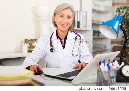 Elderly woman doctor working at laptop while sitting at table in clinic 96015066