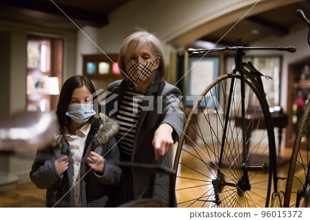 Tween schoolgirl and elderly female tutor in face masks viewing vintage bicycle in museum Tween schoolgirl and elderly female tutor in face masks viewing vintage bicycle in museum 96015372