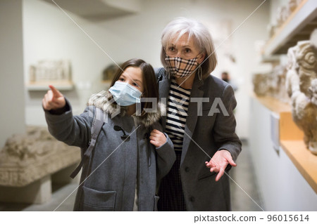 Elderly female tutor in protective mask showing tween girl ancient sculptures in museum 96015614