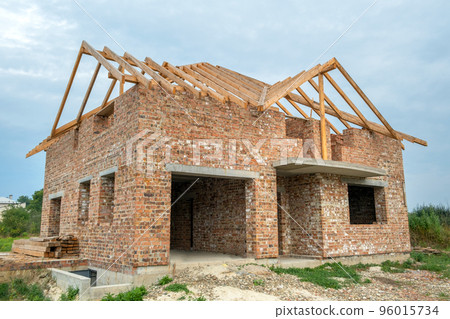 Building site with unfinished brick house with wooden roofing frame for future roof under construction. 96015734