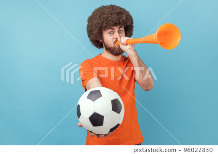 Portrait of man with Afro hairstyle wearing orange T-shirt celebrating victory of favourite football team, blowing horn and ball in hands. Indoor studio shot isolated on blue background. Portrait of man with Afro hairstyle wearing orange T-shirt celebrating victory of favourite football team, blowing horn and ball in hands. Indoor studio shot isolated on blue background. 96022830