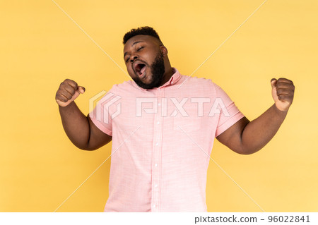 Portrait of tired bearded man wearing pink shirt yawning, feeling exhausted, lack of sleep, outstretching arms, keeps eyes closed. Indoor studio shot isolated on yellow background. Portrait of tired bearded man wearing pink shirt yawning, feeling exhausted, lack of sleep, outstretching arms, keeps eyes closed. Indoor studio shot isolated on yellow background. 96022841