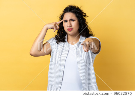 Portrait of winsome young adult woman with dark wavy hair holding finger near head and pointing finger at camera, stupid gesture. Indoor studio shot isolated on yellow background. Portrait of winsome young adult woman with dark wavy hair holding finger near head and pointing finger at camera, stupid gesture. Indoor studio shot isolated on yellow background. 96022870