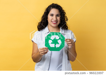 Portrait of attractive delighted woman with dark wavy hair holding green recycling sign, calls on to sort rubbish, save our planet from pollution. Indoor studio shot isolated on yellow background. 96022897