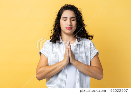 Portrait of peaceful calm relaxed woman with dark wavy hair standing with prayer gesture, keeping eyes closed, meditating sitting in yoga position. Indoor studio shot isolated on yellow background. 96022907