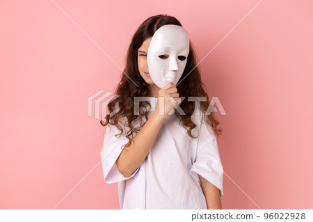 Portrait of dark haired little girl wearing white T-shirt covering face, standing with frowning face, multiple personality disorder. Indoor studio shot isolated on pink background. 96022928