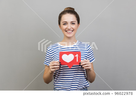 Portrait of adorable happy woman wearing striped T-shirt holding heart like icon of social media and looking at camera with toothy smile. Indoor studio shot isolated on gray background. Portrait of adorable happy woman wearing striped T-shirt holding heart like icon of social media and looking at camera with toothy smile. Indoor studio shot isolated on gray background. 96022978