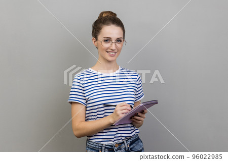 Portrait of attractive optimistic woman wearing striped T-shirt standing with paper notebook and pen in hands, looking at camera with smile. Indoor studio shot isolated on gray background. 96022985