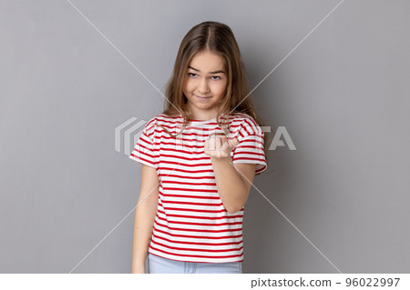 Portrait of little girl wearing striped T-shirt looking playfully and calling with one finger, making beckoning gesture, inviting to come. Indoor studio shot isolated on gray background. Portrait of little girl wearing striped T-shirt looking playfully and calling with one finger, making beckoning gesture, inviting to come. Indoor studio shot isolated on gray background. 96022997