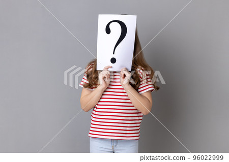 Portrait of unknown little girl wearing striped T-shirt standing hiding face behind paper with question mark, having problem for solving. Indoor studio shot isolated on gray background. 96022999