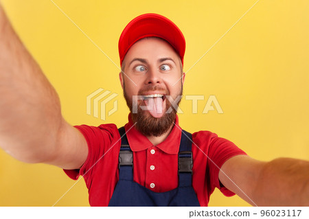 Funny handyman wearing blue uniform taking selfie, looking at camera with crossed eyes and showing tongue out POV, point of view of photo. Indoor studio shot isolated on yellow background. 96023117