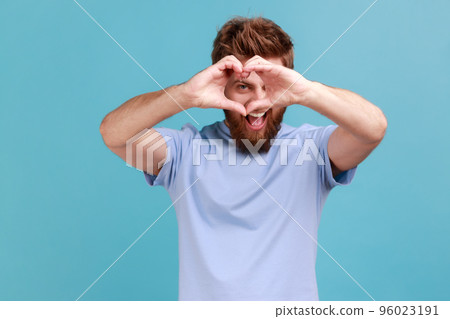 Portrait of handsome bearded man looking through hands in shape of heart showing romantic gesture, love confession, valentines day celebration. Indoor studio shot isolated on blue background. 96023191