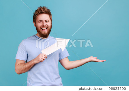Portrait of bearded man holding arrow pointing up looking at camera with toothy smile, growth and increase concept, presenting copy space. Indoor studio shot isolated on blue background. Portrait of bearded man holding arrow pointing up looking at camera with toothy smile, growth and increase concept, presenting copy space. Indoor studio shot isolated on blue background. 96023200