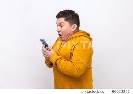 Side view of man using mobile phone with surprised expression, addicted to smartphone, texting in social network, wearing urban style hoodie. Indoor studio shot isolated on white background. 96023201