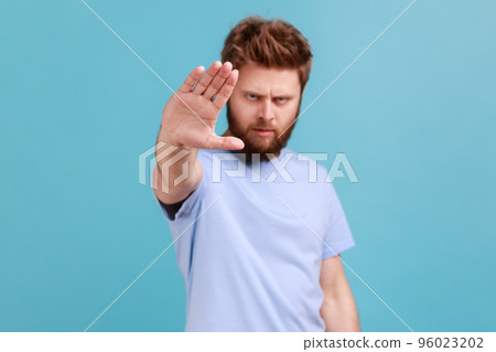 Portrait of strict bearded man showing stop sign at camera, standing with frowning face and bossy expression, meaning caution to avoid danger or mistake. Indoor studio shot isolated on blue background 96023202