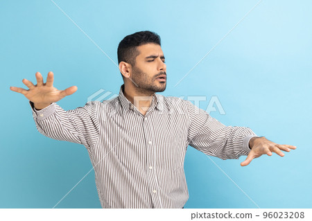 Loneless and blindness. Lost worry bearded businessman standing with closed eyes and try to touching something or find, wearing striped shirt. Indoor studio shot isolated on blue background. Loneless and blindness. Lost worry bearded businessman standing with closed eyes and try to touching something or find, wearing striped shirt. Indoor studio shot isolated on blue background. 96023208