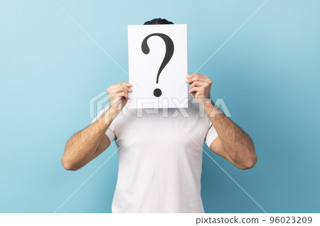 Portrait of man wearing white T-shirt hiding his face behind white paper with question mark, finding smart solution, asking for advice. Indoor studio shot isolated on blue background. Portrait of man wearing white T-shirt hiding his face behind white paper with question mark, finding smart solution, asking for advice. Indoor studio shot isolated on blue background. 96023209