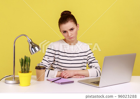 Bored unhappy woman office manager wearing striped shirt sitting at workplace looking at camera, tired of online conference. Indoor studio studio shot isolated on yellow background. Bored unhappy woman office manager wearing striped shirt sitting at workplace looking at camera, tired of online conference. Indoor studio studio shot isolated on yellow background. 96023288