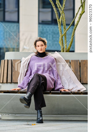 Young woman in wide trousers, sweater, coat and rough boots sits on bench Young woman in wide trousers, sweater, coat and rough boots sits on bench 96023370