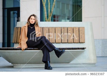 Young woman in wide trousers, sweater, suede jacket and rough boots sits on bench Young woman in wide trousers, sweater, suede jacket and rough boots sits on bench 96023387