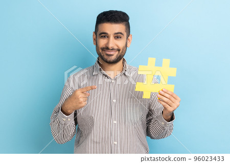 Viral hashtag and successful blogging. Smiling businessman with beard pointing at large yellow hash symbol, wearing striped shirt. Indoor studio shot isolated on blue background. Viral hashtag and successful blogging. Smiling businessman with beard pointing at large yellow hash symbol, wearing striped shirt. Indoor studio shot isolated on blue background. 96023433