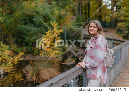 Portrait Of Cute Young Woman In Casual Wear In Autumn, Standing On Bridge Against Background Of An Autumn Park And River. Pretty Female Walking In Park In Golden Fall. Copy Space. Smiling Girl In The 96024345