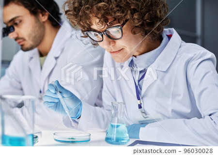 Young clinician adding some drops of fluid in pipette into petri dish with bacteria while carrying out scientific experiment with colleague 96030028
