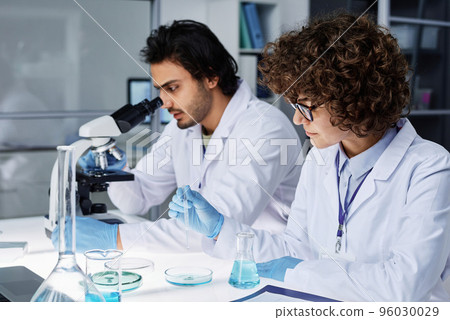 Young clinicians working in team over new scientific experiment while man looking in microscope and woman mixing liquid substances 96030029