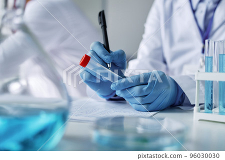 Close-up of gloved hands of clinician with pen writing down sample name on test tube with new vaccine or chemical substance 96030030
