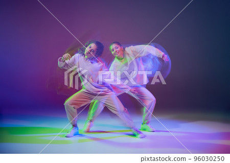 Portrait of young girls dancing hip-hop isolated over gradient blue purple background in neon with mixed light. Multicolored 96030250