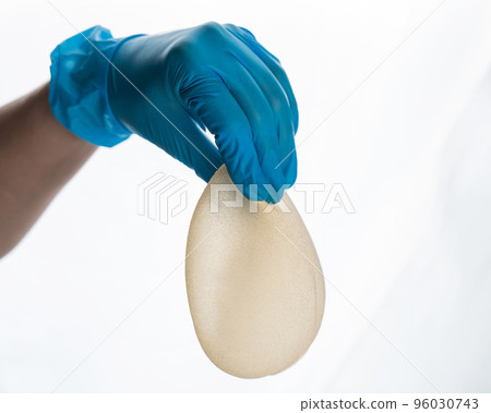 A female hand in gloves holds a teardrop-shaped breast implant on a white background. A female hand in gloves holds a teardrop-shaped breast implant on a white background. 96030743