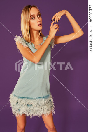 Image of cute beautiful slim blonde woman in elegant cocktail blue dress is posing at studio on purple background. Image of cute beautiful slim blonde woman in elegant cocktail blue dress is posing at studio on purple background. 96031572