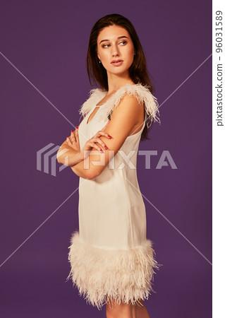 Beautiful brunette woman in elegant cocktail white dress is posing in studio on purple background. Beautiful brunette woman in elegant cocktail white dress is posing in studio on purple background. 96031589