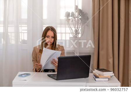 European professional woman sitting with laptop at home office desk, positive woman studying while working on PC. She is wearing a beige jacket and jeans. 96032757