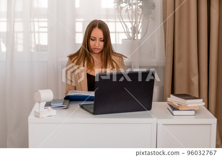 European professional woman sitting with laptop at home office desk, positive woman studying while working on PC. She is wearing a beige jacket and jeans. European professional woman sitting with laptop at home office desk, positive woman studying while working on PC. She is wearing a beige jacket and jeans. 96032767