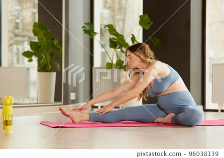 Portrait of young pregnant woman sitting on mat and stretching, doing yoga indoors on a daytime Portrait of young pregnant woman sitting on mat and stretching, doing yoga indoors on a daytime 96038139