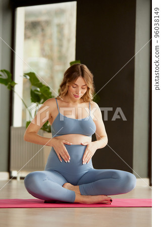 Portrait of young pregnant woman in sportswear sitting on yoga mat and holding belly with heart shape. 96038149