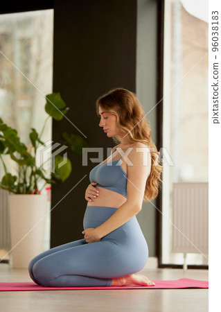 Portrait of young pregnant woman doing yoga, sitting on mat and holding belly indoors on a daytime. Meditation 96038183