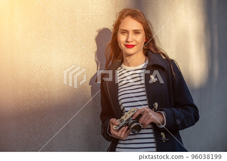 Smiling female photographer in jacket standing in front of wall ready to make new photo. Sun flare Smiling female photographer in jacket standing in front of wall ready to make new photo. Sun flare 96038199