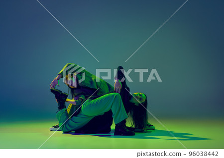 Young stylish flexible girls in modern attires dancing contemp style dance isolated over gradient blue-green background at dance hall in neon light. Young stylish flexible girls in modern attires dancing contemp style dance isolated over gradient blue-green background at dance hall in neon light. 96038442
