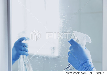 Cropped view of housekeeper woman hand in rubber blue gloves spraying chemical cleaner liquid to the mirror while cleaning bathroom at home. Housekeeping and cleaning service. Clean house, cleanliness 96038974