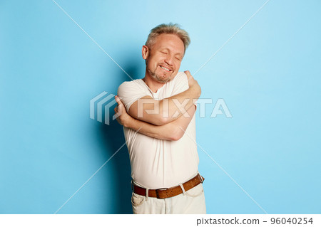 Portrait of mature man in white T-shirt posing, hugging himself isolated over blue studio background Portrait of mature man in white T-shirt posing, hugging himself isolated over blue studio background 96040254