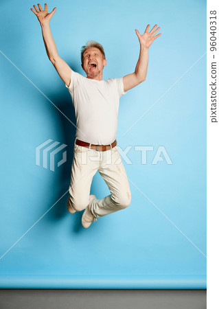 Portrait of mature man in white casual clothes posing, cheerfully jumping isolated over blue studio background Portrait of mature man in white casual clothes posing, cheerfully jumping isolated over blue studio background 96040318
