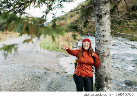 Happy redhead Woman in red hat hiking in woods. Adventure women standing near the river and pointing finger on mountains background. Freedom and active lifestyle concept. Amazing chill moment Happy redhead Woman in red hat hiking in woods. Adventure women standing near the river and pointing finger on mountains background. Freedom and active lifestyle concept. Amazing chill moment 96040706