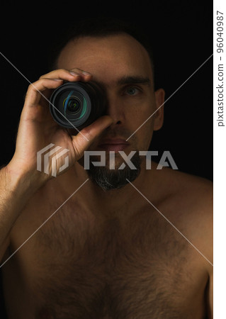 Close-up of a man with a lens leaning against his eye. Black background. Shirtless photographer using lens instead of eye. Breast portrait of a Caucasian bearded man. Low key. Man looking into camera. Close-up of a man with a lens leaning against his eye. Black background. Shirtless photographer using lens instead of eye. Breast portrait of a Caucasian bearded man. Low key. Man looking into camera. 96040987