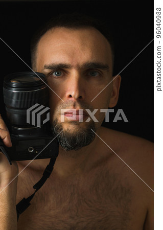 Portrait of photographer on black background. Shirtless bearded man holding camera near face with lens up. Low key. Portrait of photographer on black background. Shirtless bearded man holding camera near face with lens up. Low key. 96040988