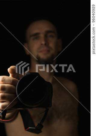 Shirtless man holding the camera at arm's length. Belt portrait of a photographer on a black background. Focus on the camera. Low key. Selective focus. Shirtless man holding the camera at arm's length. Belt portrait of a photographer on a black background. Focus on the camera. Low key. Selective focus. 96040989