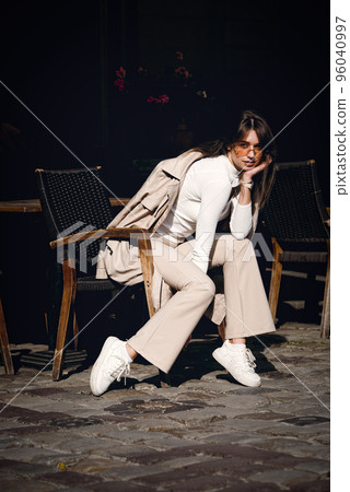 beautiful brunette girl dressed in white blouse, pants, sneakers with a handbag and camel coat in hands. Stylish trendy fashion outlook 96040997