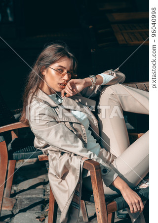 beautiful brunette girl in a sunglasses dressed in white blouse and beige coat posing in a chair. Stylish trendy fashion outlook 96040998